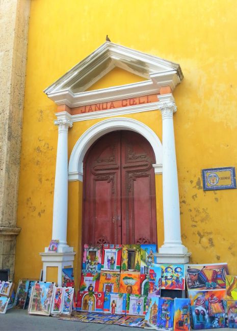 Colourful Cartagena Yellow Church with Arched Doorway and colourful artwork paintings propped up for sale in the doorway- Solo Travel in Colombia