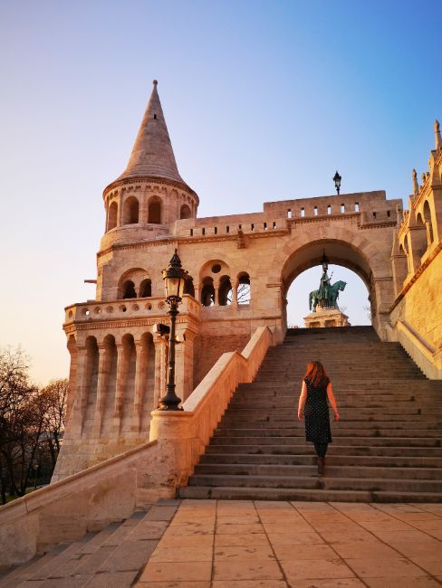 Solo Female Travel in Budapest - Hungary - Claire walking up steps away from camera to a turret and statue in an archway at the top of the steps